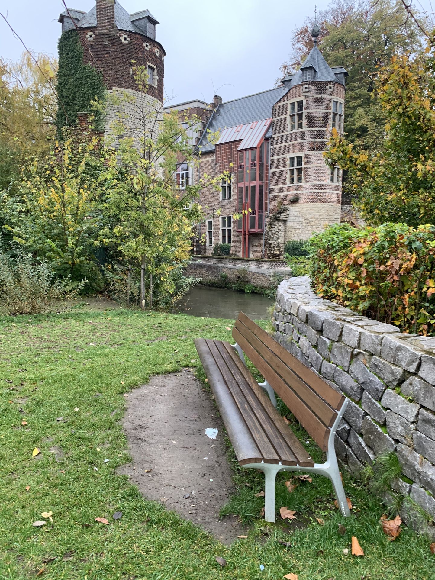 Een klassiek ogende, duurzame zitbank met rugleuning uit de NeoRomántico Clásico-serie van Urbidermis, geplaatst in een groene, parkachtige omgeving in het Janseniuspark in Leuven. De bank, met latten van tropisch hardhout in een natuurlijke kleur, contrasteert met de lichtgrijze, sierlijke metalen onderstellen. De bank staat op de rand van een klein grindpad, grenzend aan een stenen muur en een waterloop, perfect geïntegreerd in de historische en groene context van het park.