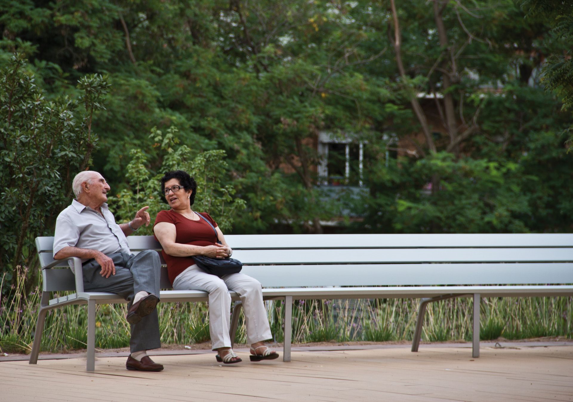 Urbidermis Neoromantico Liviano Aluminio Oudere man en vrouw ontspannen op een ergonomische zitbank in een groene openbare ruimte, bevordert inclusie en toegankelijkheid.