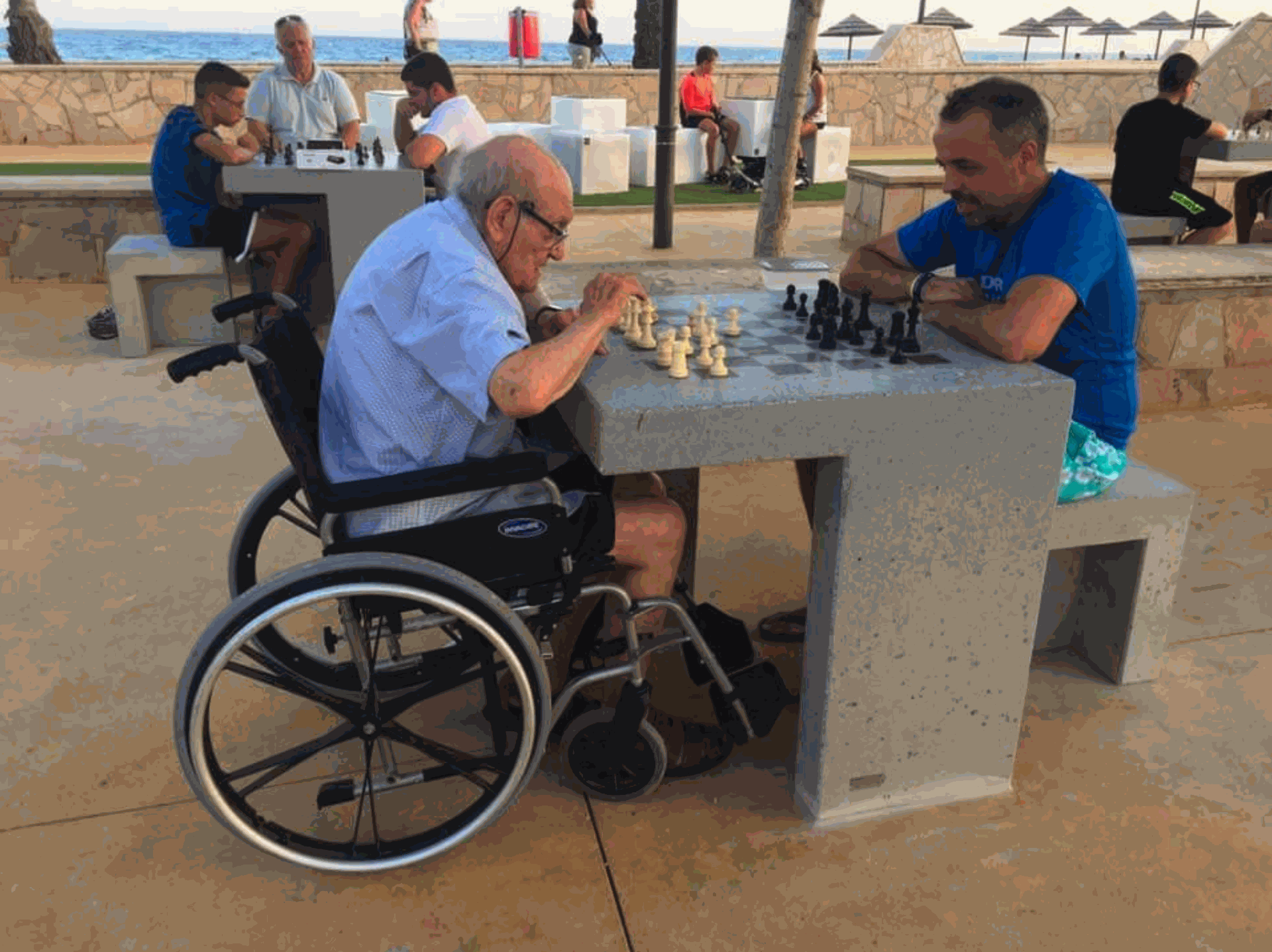 Escofet Prat Mesa Chess Man in een rolstoel speelt schaak aan een rolstoelvriendelijke tafel in een park aan zee, voorbeeld van inclusieve recreatie.