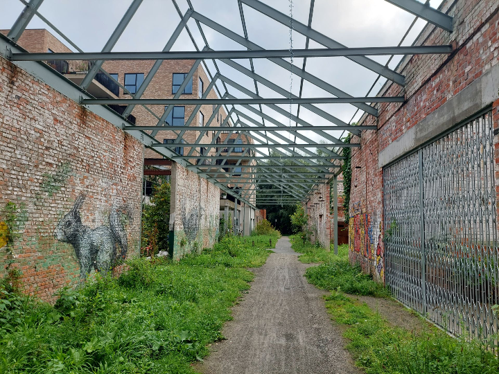 Groene doorgang in Bijgaardepark, Gent, met straatkunst van ROA op bewaarde bakstenen muren en een open stalen structuur als herinnering aan het industriële verleden.