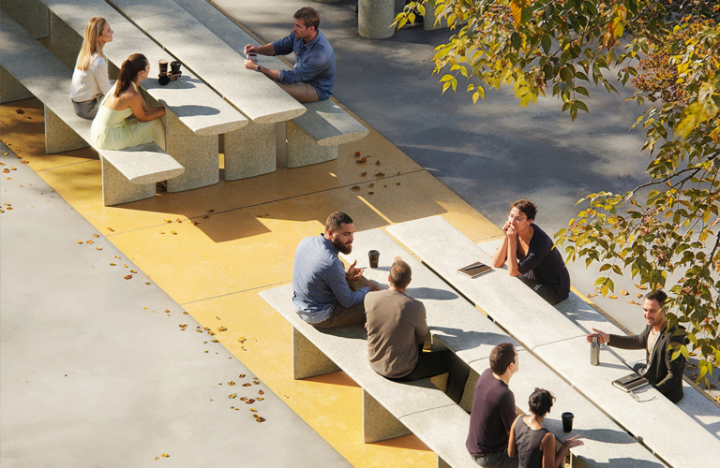 Bancs et tables modulaires en béton de la collection Tiers d’Escofet sur une place urbaine colorée, avec des utilisateurs en pleine discussion sous les arbres d’automne.