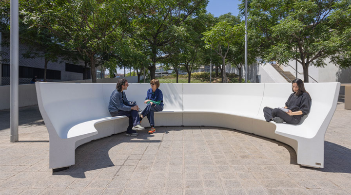 Banc courbé modulaire Gather d’Escofet en demi-cercle, utilisé comme lieu de rencontre dans un espace urbain ombragé.