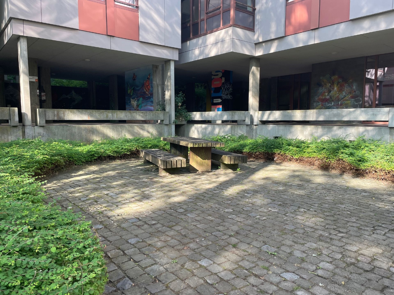 Een robuuste houten tafel met bijpassende banken zonder rugleuning staat op een kasseienterras, omringd door groen en moderne studentenresidenties op de campus in Leuven.