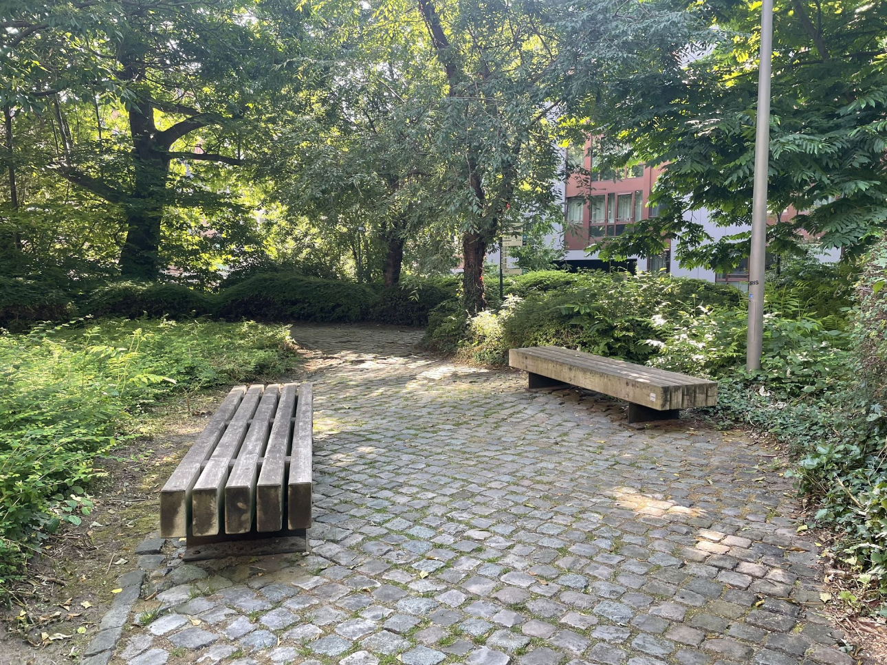 Twee houten Tramet zitbanken zonder rugleuning staan in een groene, klimaatbestendige binnentuin met kasseien op de campus van studentenresidenties in Leuven.