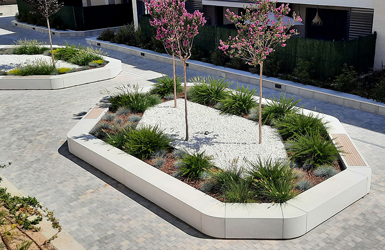 Boxland : île verte modulaire qui rend le parcours et le séjour lisibles. Grand îlot périmétrique en béton clair avec assise, végétation stratifiée et deux arbres en rue apaisée.