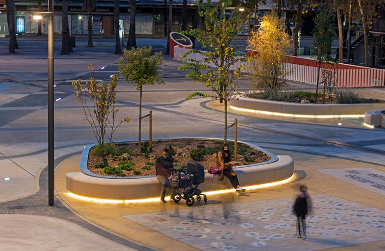 Comu zitbanken van Escofet in zachte aardetinten op een open buurtplein, met gras en bomen, ontworpen voor comfort, rust en samenhang.