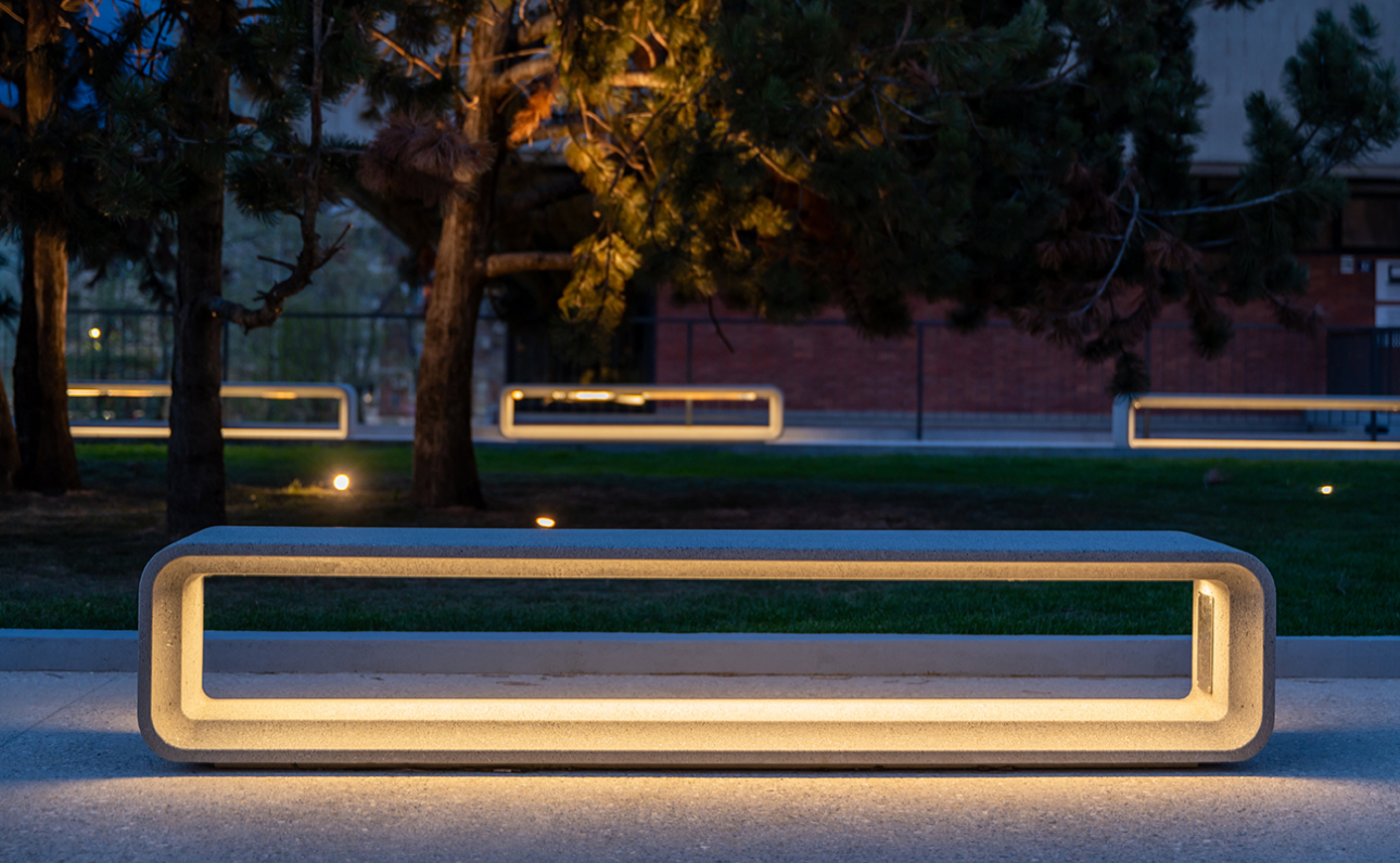 Mom zitbanken van Escofet met geïntegreerde LED-verlichting rondom beplanting op een stedelijk plein, voor zichtbaarheid en sfeer na zonsondergang.