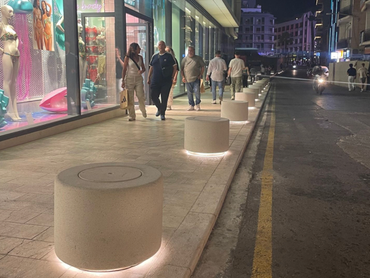 Winkelstraat bij nacht met brede stoep en rij cilindervormige betonnen zitelementen met zachte LED-verlichting die stoep en rijweg scheiden.