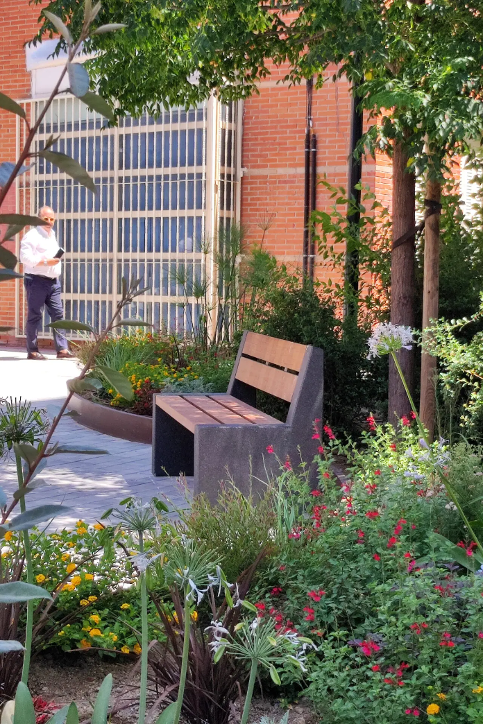 Alpine buitenbank in groen aangelegde binnentuin bij gebouw, betonnen steunen en houten latten als rustpunt in stedelijke setting