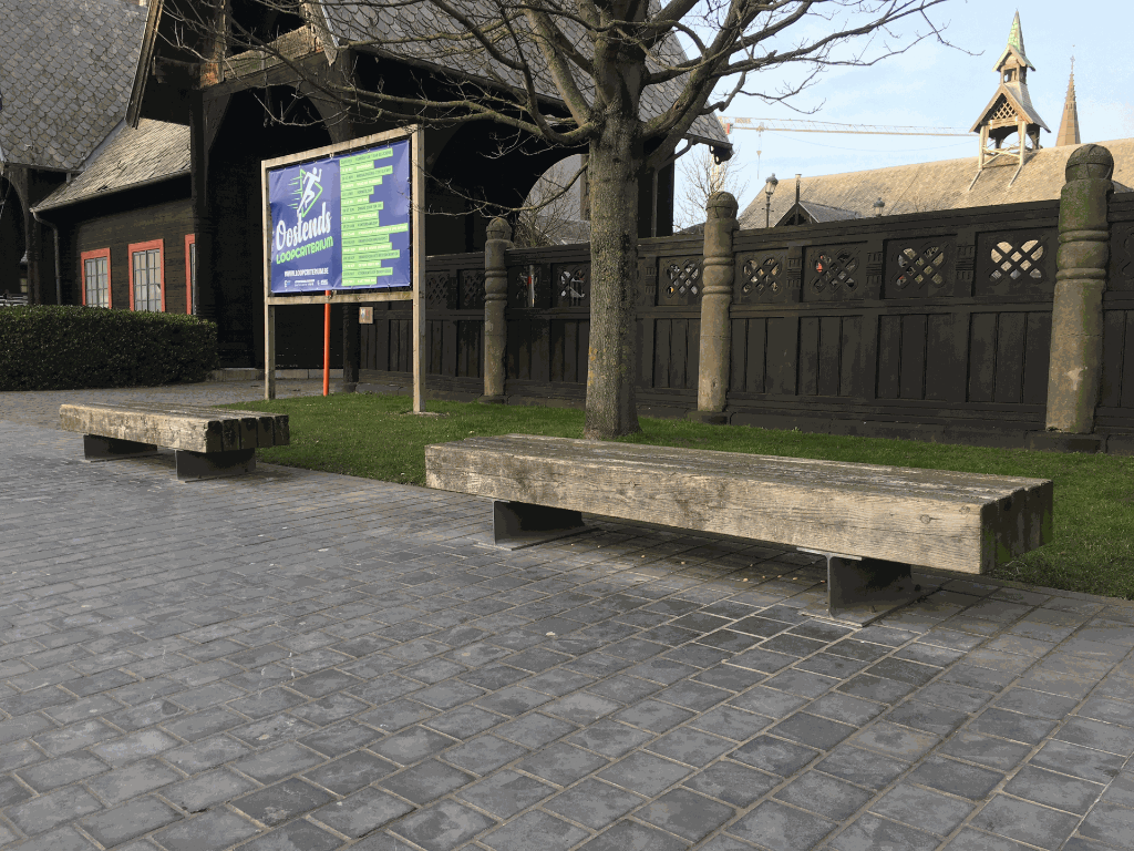 De Tramet zitbanken van Escofet 1886 in Oostende; een robuuste houten zitbanken op stalen voeten langs een verharde wandelzone bij een historische site met donkere houten omheining, bomen en erfgoedarchitectuur.