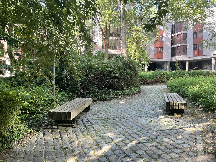 Houten zitbank Tramet op stalen onderstel in een groene binnentuin tussen appartementsgebouwen in Camillo Torres te Leuven, geplaatst langs een waterdoorlatend pad met kasseien en weelderige beplanting