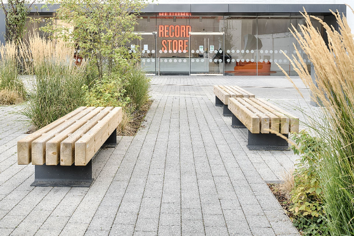 Twee robuuste houten zitbanken op stalen onderstel langs een brede wandelzone met grijze bestrating, siergrassen en vaste beplanting aan de ingang van een openbaar gebouw.