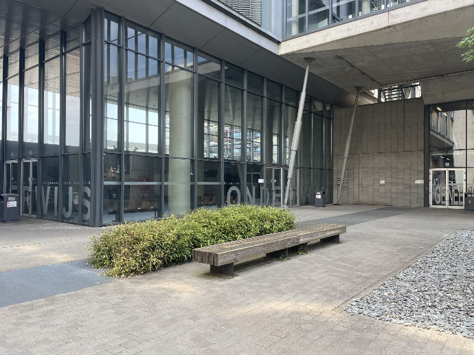 Lange houten zitbank op een lage stalen constructie naast een groen plantvak voor een modern openbaar gebouw met glasgevel en betonnen architectuur op de Campus Gasthuisberg in Leuven