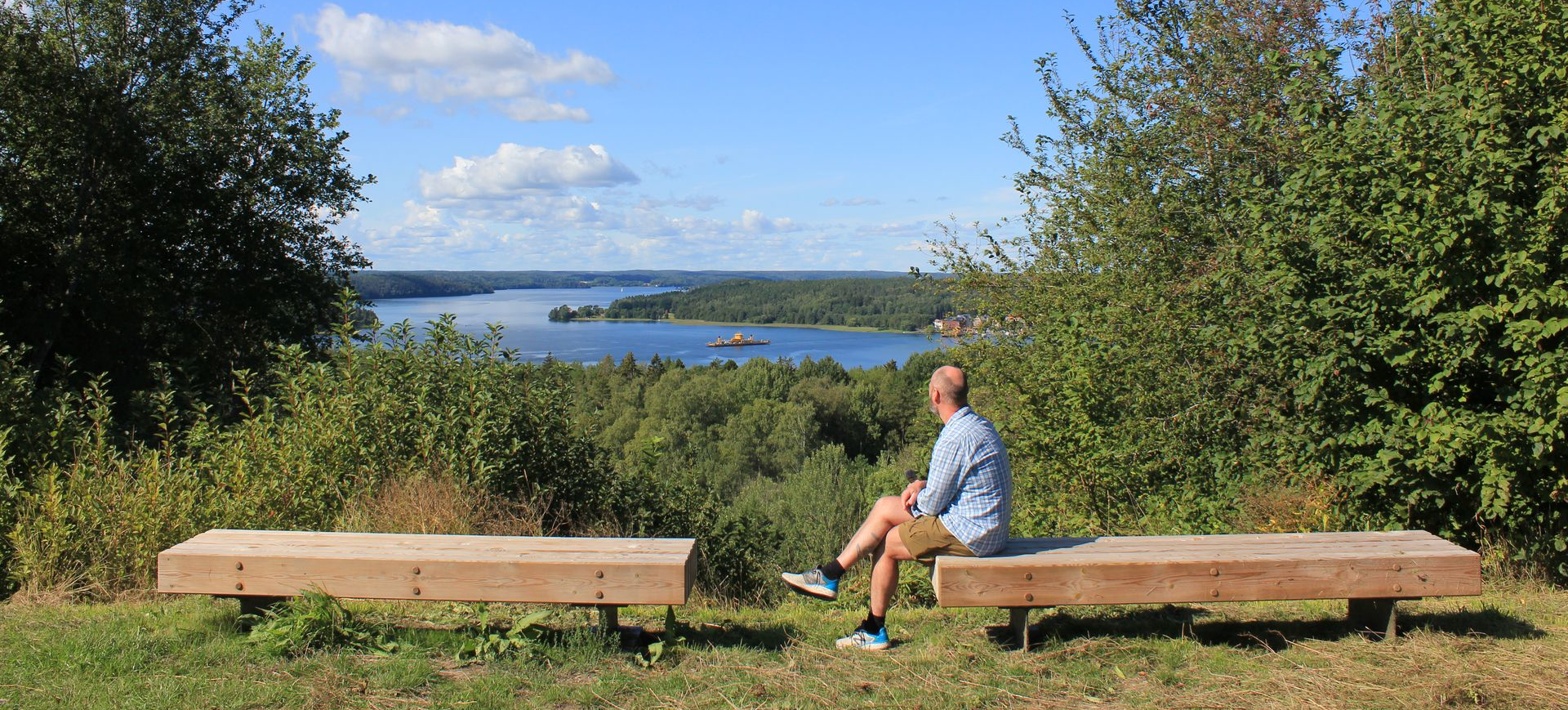 Twee massieve houten banken op een uitkijkpunt in een groen natuurgebied, met zicht op een meer, bossen en een open landschap onder een blauwe hemel.