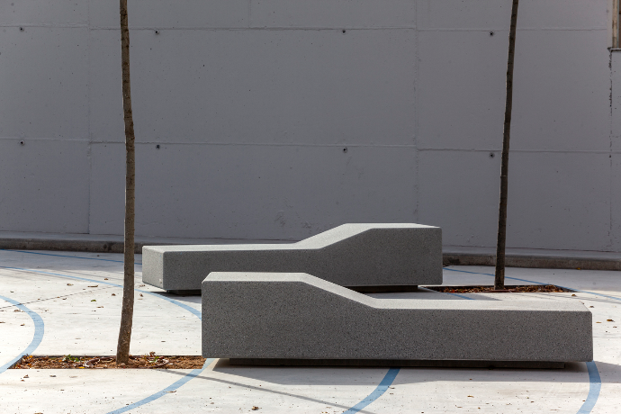 Twee Garonne Aran zitbanken van Escofet 1886 in grijs beton, met verhoogd profiel en rugloze chaise longue vorm tussen jonge bomen op een stedelijk sport- en speelplein.