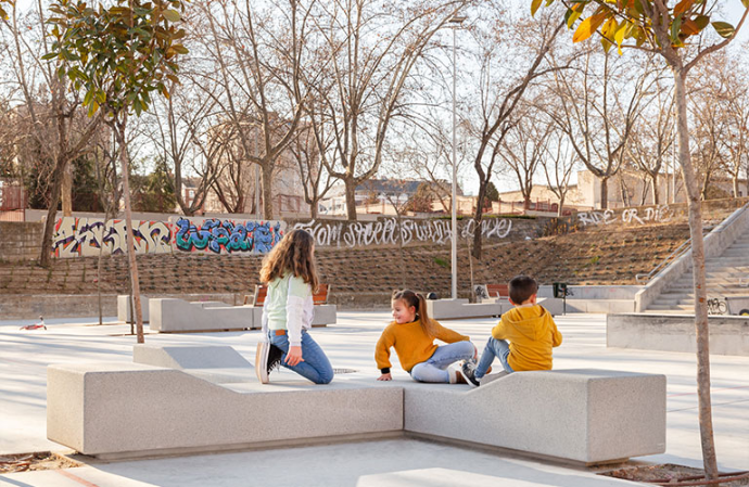 Garonne modulaire zitbank van Escofet 1886 in licht beton op een stedelijk speelplein, met rugloze chaise longue vorm en afgeronde randen voor flexibel zitten, spelen en ontmoeten.